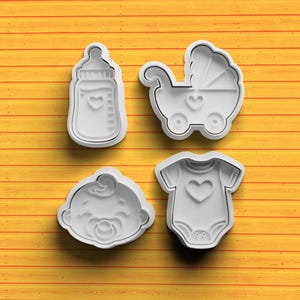Può includere: Quattro formine bianche di plastica a tema baby. Le forme includono un biberon, una carrozzina, un viso di bambino e un body. Ogni formina ha una forma di cuore in rilievo. Le formine sono su una superficie di legno giallo.
