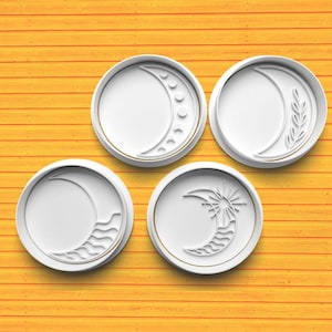Pode incluir: Quatro cortadores de biscoitos circulares brancos com borda dourada, cada um com um design de lua crescente diferente. Um tem uma série de pequenos círculos, outro um galho, um terceiro linhas onduladas e o último um sol.