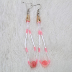 May include: A pair of dangle earrings with a gold-colored cone top, white and pink beaded strands, and a large pink bead at the bottom. The earrings are displayed on a white, fluffy surface.