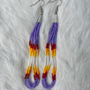 May include: A pair of beaded earrings with silver-tone hooks. The earrings feature strands of purple, red, yellow, and white beads. The beads are arranged in a vertical pattern, with a loop at the bottom.