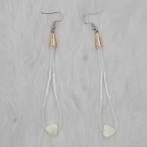 May include: A pair of dangle earrings with gold-colored cone-shaped tops and silver-colored fish hook closures. The earrings feature long strands of small white beads, culminating in a heart-shaped charm at the bottom.