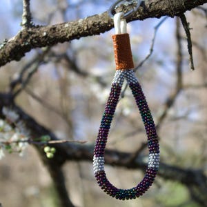 May include: A beaded keychain with a loop design, hanging from a tree branch. The keychain features a brown leather accent, a silver-gray section, and a loop of dark purple, green, and black beads. The background is blurred with tree branches and blossoms.