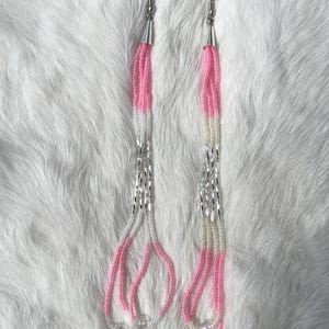 May include: A pair of dangle earrings with pink and white beaded strands. The earrings have silver-tone metal hooks and accents. The beaded strands are arranged in a cascading design, creating a delicate and feminine look. The earrings are displayed on a white, textured surface.
