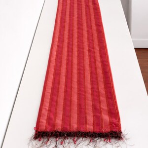 Pode incluir: Um caminho de mesa listrado em vermelho e rosa com borda de franjas. O caminho de mesa é feito de um tecido transparente e possui listras verticais em tons variados de vermelho e rosa. A borda de franjas adiciona um toque decorativo.
