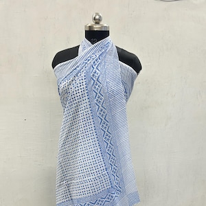 May include: A white and blue patterned sarong is draped over a mannequin. The sarong features a geometric design with blue stripes and a border with a repeating pattern. The fabric is lightweight and appears to be cotton.