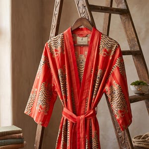 Könnte beinhalten: Ein leuchtend roter Kimono-Bademantel mit einem sich wiederholenden Muster aus schwarzen und beigen Tigern. Das Kleidungsstück hat lange Ärmel, einen breiten Kragen und einen passenden Stoffgürtel in der Taille. Ein Etikett im Nacken zeigt PAAKSH.