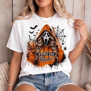 May include: A white short-sleeve t-shirt featuring a graphic of a figure in an orange hoodie with a ghost mask, holding a pumpkin-faced drink. The design includes bats, a spider web, and the text "HALLOWEEN Mama" in black, with orange paint drips.