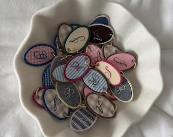 May include: A collection of embroidered oval tags with various initial letters in a white, scalloped bowl. The tags feature different patterns, including stripes and gingham, with gold-colored metal eyelets.