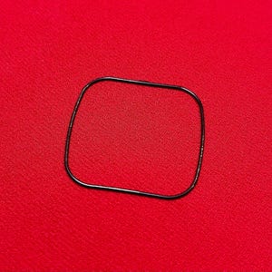 May include: A thin, black, rectangular rubber gasket sits on a red surface. The gasket has rounded corners and appears to be made of a flexible material. The background is a solid, vibrant red.
