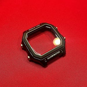 May include: A black watch case with a clear screen, featuring the text "WORLD TIME" and "ILLUMINATOR". The watch case is set against a red background. The watch case has a square shape with rounded corners and small buttons on the sides.