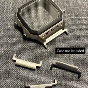 Puede incluir: Una caja de reloj de metal plateado con un bisel negro y el texto WORLD TIME impreso en la parte frontal. Tres adaptadores de correa de metal se encuentran debajo de la caja. Una etiqueta negra muestra el texto Case not included.
