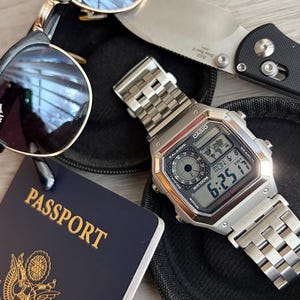 May include: A silver-tone Casio digital watch with a metal link band, black-rimmed Ray-Ban sunglasses, a folding pocket knife with a black handle, and a dark blue United States passport featuring a gold eagle emblem on the front cover.