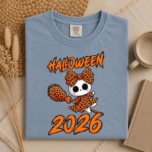 Peut inclure: Un t-shirt bleu avec un motif représentant un fantôme blanc portant un nœud et une robe à imprimé léopard, tenant un balai assorti. Le texte "HALOWEEN" est inscrit au-dessus et "2026" en dessous en lettres orange vif.
