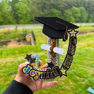 Puede incluir: Una decoración personalizada con la letra J que incluye un birrete de graduación negro con purpurina, un diploma blanco enrollado y el nombre Joyniel en letras azules y doradas. La letra tiene un acabado holográfico plateado con estrellas.