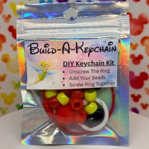 Puede incluir: Un kit de llavero DIY en una bolsa transparente e iridiscente. El kit incluye cuentas rojas, amarillas y negras, un anillo blanco y un cordón rojo. El paquete dice "Build-A-Keychain" y "DIY Keychain Kit".