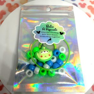 May include: A DIY keychain kit in a holographic pouch containing a green alien character charm, a lime green wire ring, and assorted blue, green, and white plastic beads. The label reads: Build A Keychain, Unscrew The Ring, Add Your Beads, Screw Ring Together.