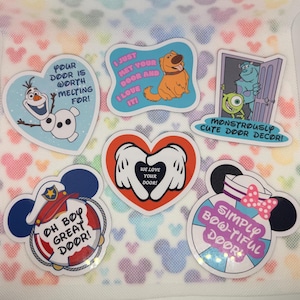 Puede incluir: Una colección de seis pegatinas decorativas con personajes de dibujos animados y texto. Los diseños incluyen a Olaf con 'Your door is worth melting for!', Dug con 'I just met your door and I love it!', Mike y Sulley, guantes blancos formando un corazón con 'We love your door!', un sombrero de marinero con 'Oh boy great door!' y un lazo con 'Simply bow-tiful door!'