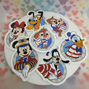 May include: A selection of colourful Disney character stickers featuring Mickey Mouse, Minnie Mouse, Donald Duck, Pluto, Goofy, Chip, and Dale. Each sticker shows a character in a circular frame with a nautical theme. The stickers are on a white surface.