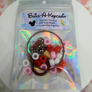 Può includere: Una busta di plastica trasparente contenente un kit Build-A-Keychain. Il kit include un portachiavi marrone, perline colorate rosa, bianche e rosse e un ciondolo a forma di testa di Topolino. Il testo "Build-A-Keychain" è stampato sull'etichetta.