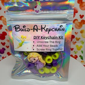 May include: A DIY keychain kit in a holographic bag. The kit includes a small doll, purple and yellow beads, and a keychain ring. The bag has the text "Build-A-Keychain" and instructions.