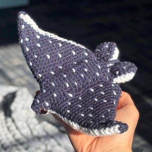 K&ouml;nnte beinhalten: Ein handgeh&auml;kelter, dunkelblauer Walhai mit wei&szlig;en Punkten und wei&szlig;er Bord&uuml;re. Das Pl&uuml;schtier hat kleine Flossen und ein Knopfauge. Der Walhai wird in einer Hand gehalten.