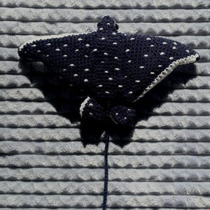 K&ouml;nnte beinhalten: Eine handgefertigte, dunkelblaue geh&auml;kelte Mantarochen mit wei&szlig;en Punkten und einem wei&szlig;en Rand. Der Rochen hat zwei kleine Flossen und einen langen, dunkelblauen Schwanz. Der Artikel wird auf einer grauen, strukturierten Oberfl&auml;che pr&auml;sentiert.