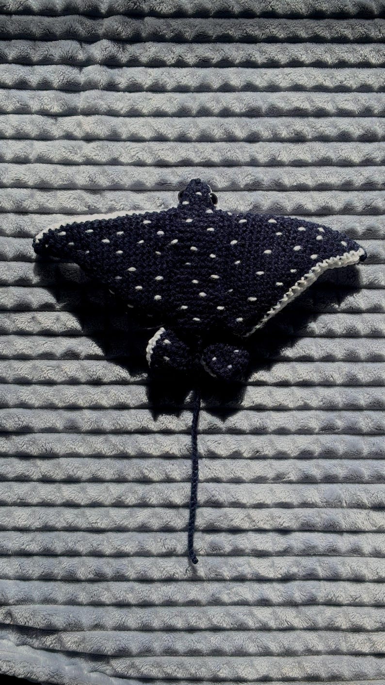 K&ouml;nnte beinhalten: Eine handgefertigte, dunkelblaue geh&auml;kelte Mantarochen mit wei&szlig;en Punkten und einem wei&szlig;en Rand. Der Rochen hat zwei kleine Flossen und einen langen, dunkelblauen Schwanz. Der Artikel wird auf einer grauen, strukturierten Oberfl&auml;che pr&auml;sentiert.
