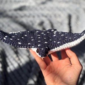 K&ouml;nnte beinhalten: Ein handgefertigtes, geh&auml;keltes Rochenspielzeug in Marineblau mit wei&szlig;en Punkten und einem wei&szlig;en Rand. Das Spielzeug hat kleine, schwarze Augen und wird in einer Hand gehalten. Der Hintergrund ist ein weicher, strukturierter Stoff.