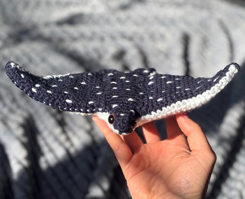 K&ouml;nnte beinhalten: Ein handgefertigtes, geh&auml;keltes Rochenspielzeug in Marineblau mit wei&szlig;en Punkten und einem wei&szlig;en Rand. Das Spielzeug hat kleine, schwarze Augen und wird in einer Hand gehalten. Der Hintergrund ist ein weicher, strukturierter Stoff.