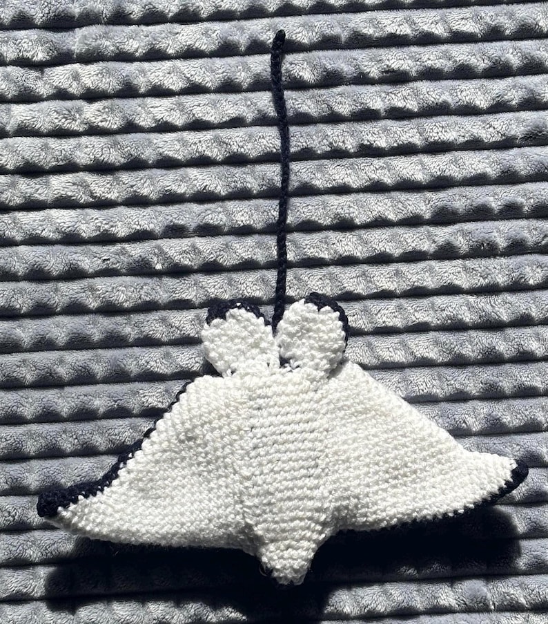 K&ouml;nnte beinhalten: Handgefertigter, wei&szlig;-blauer Strickrochen-Ornament. Der Rochen hat einen flachen, rautenf&ouml;rmigen K&ouml;rper mit abgerundeten Kanten und einem langen, d&uuml;nnen Schwanz. Die Kanten und der Schwanz sind marineblau, der K&ouml;rper ist wei&szlig;. Das Ornament wird von einer marineblauen Schnur gehalten.