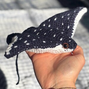 K&ouml;nnte beinhalten: Handgefertigtes H&auml;keltier in Form eines Mantarochens. Das Spielzeug ist marineblau mit wei&szlig;en Punkten und einem wei&szlig;en Rand. Es hat einen kleinen schwarzen Schwanz und braune Augen. Das Spielzeug ist aus Garn gefertigt und ein weiches Pl&uuml;schtier.
