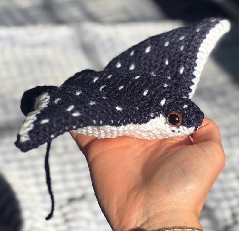 K&ouml;nnte beinhalten: Handgefertigtes H&auml;keltier in Form eines Mantarochens. Das Spielzeug ist marineblau mit wei&szlig;en Punkten und einem wei&szlig;en Rand. Es hat einen kleinen schwarzen Schwanz und braune Augen. Das Spielzeug ist aus Garn gefertigt und ein weiches Pl&uuml;schtier.