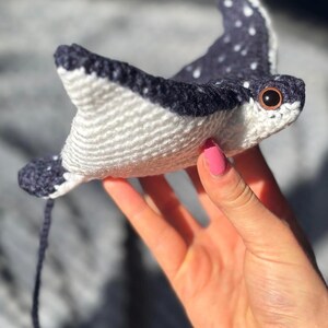 K&ouml;nnte beinhalten: Handgefertigtes H&auml;keltier in Form eines Mantarochens. Das Spielzeug hat einen wei&szlig;en K&ouml;rper mit dunkelblauen Fl&uuml;geln, die mit wei&szlig;en Punkten versehen sind. Der Rochen hat braune Augen und einen dunkelblauen Schwanz. Das Spielzeug wird in einer Hand gehalten.