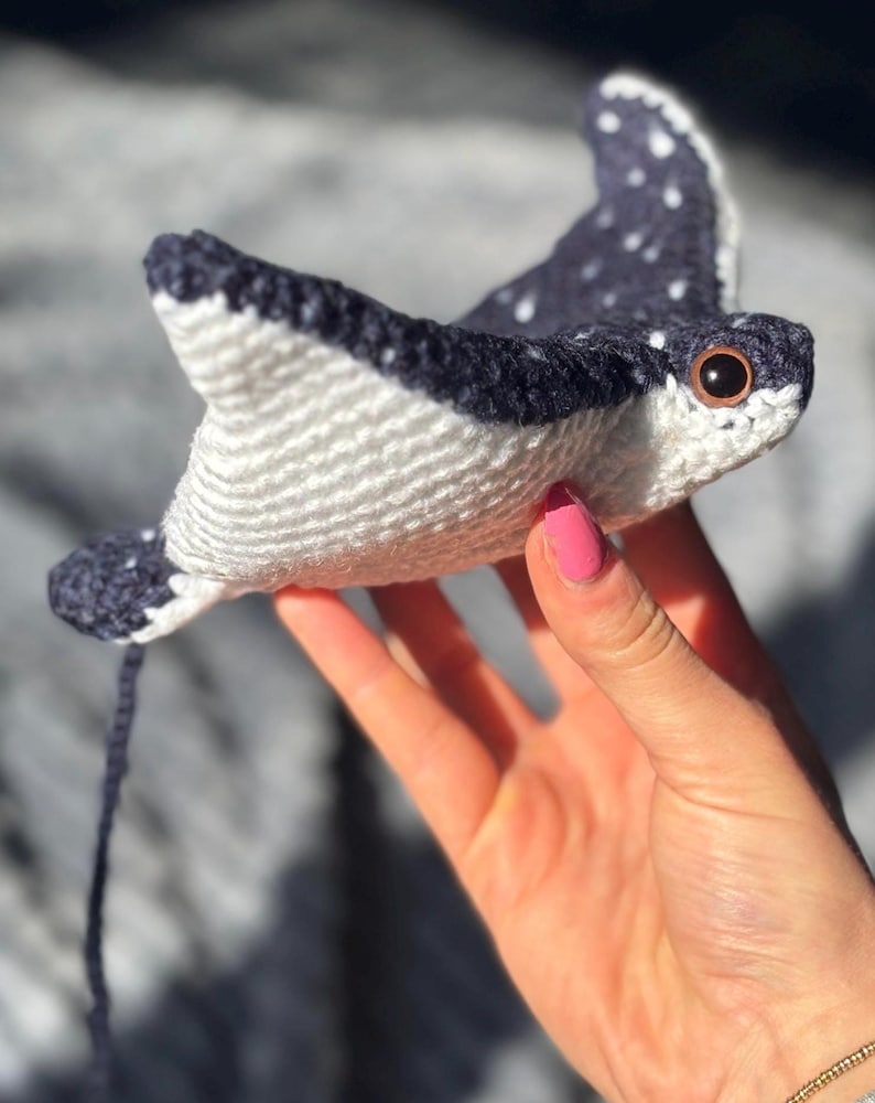 K&ouml;nnte beinhalten: Handgefertigtes H&auml;keltier in Form eines Mantarochens. Das Spielzeug hat einen wei&szlig;en K&ouml;rper mit dunkelblauen Fl&uuml;geln, die mit wei&szlig;en Punkten versehen sind. Der Rochen hat braune Augen und einen dunkelblauen Schwanz. Das Spielzeug wird in einer Hand gehalten.