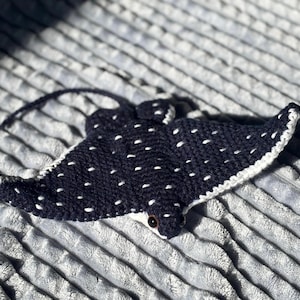 May include: A handmade, dark blue crochet stingray with white polka dots and a white border. The stingray has a small, dark eye and a long, thin tail. The craft is lying on a textured, gray surface.