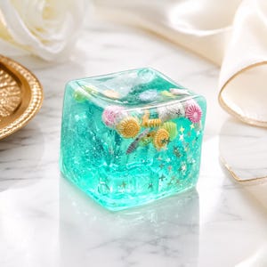 Peut inclure: Un cube presse-papier en résine bleu sarcelle translucide contenant des coquillages miniatures incrustés dans des tons rose, jaune et blanc, ainsi que de petites paillettes dorées en forme d'étoile suspendues dans le bloc carré brillant.