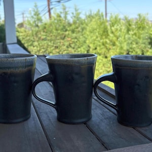 May include: Three ceramic mugs with a dark charcoal finish and a subtle, lighter-toned rim. Each mug features a curved handle and a slightly tapered cylindrical body. The surface of the mugs has a smooth, matte texture with visible variations in color.