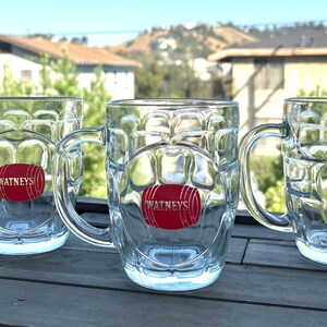 May include: Three clear glass beer mugs with a dimpled texture and a sturdy handle. Each mug features a red oval logo on the front with a graphic of a barrel and the text WATNEYS printed in white capital letters across the center of the barrel.