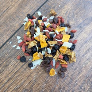 May include: A mixture of dried food ingredients including bright orange pieces of dried mango, dark purple raisins, small red goji berries, and thin white flakes of coconut. The assortment of dried fruits is scattered in a loose pile.