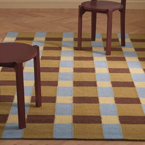 Puede incluir: Una alfombra rectangular con un patrón de cuadrícula repetitivo en tonos azul claro, amarillo mostaza y marrón oscuro. Dos taburetes redondos de color granate oscuro con cuatro patas rectas están colocados sobre la alfombra, uno al frente y otro detrás.