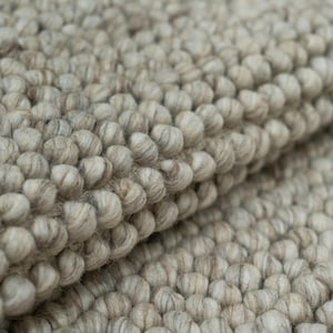 Può includere: Un tappeto spesso e strutturato caratterizzato da una densa costruzione annodata a mano di palline di lana infeltrita. La superficie presenta una tavolozza di colori variegati nei toni del beige tenue, grigio chiaro e crema, simile a ciottoli.