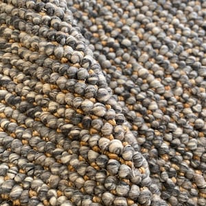 May include: Close-up of a textured rug with a looped pile. The rug features a mix of grey, white, and tan yarns, creating a speckled appearance. The fibres are tightly woven, and the rug has a thick, durable construction.