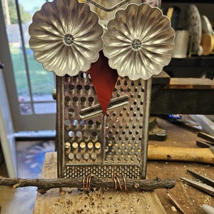 May include: A handcrafted owl sculpture made from a repurposed cheese grater. The owl features silver flower-shaped eyes, a red felt beak, and copper wire feet perched on a small branch. The body is the grater, and the sculpture is on a wooden surface.
