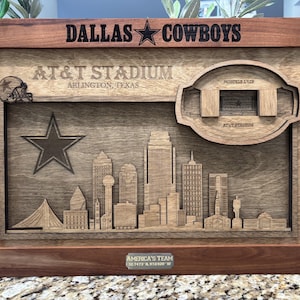 May include: Wooden framed artwork featuring the Dallas Cowboys logo and the text "DALLAS COWBOYS". The artwork includes a detailed depiction of AT&T Stadium in Arlington, Texas, and a cityscape. The words "AMERICA'S TEAM" are also visible.