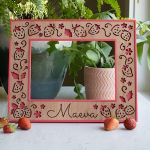 May include: A wooden picture frame with a pink border and strawberry cutouts. The name "Maeva" is engraved at the bottom. Fresh strawberries are scattered in front of the frame. The frame is displayed near a window with plants.