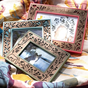 May include: Three decorative wooden picture frames with floral and bear cutouts. One frame has a pink border, another has a light blue border, and the third has a green border. The frames are on a colorful floral patterned fabric.