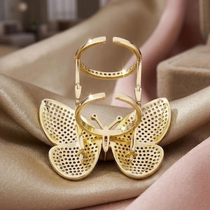 May include: A gold-colored butterfly ring with a unique design. The ring features a butterfly-shaped base with intricate cut-out details and a double-banded ring design. The ring is set on a satin surface.