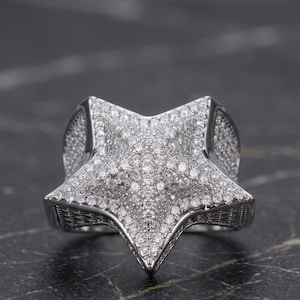 May include: A silver-toned ring in the shape of a star, encrusted with numerous small, sparkling stones. The ring has a three-dimensional design with pointed edges and a textured band.