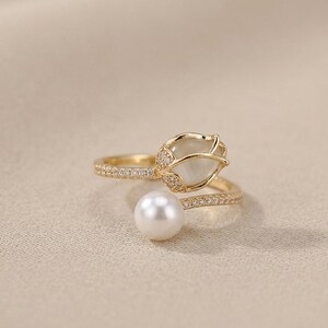 May include: A gold-colored ring with a floral design, featuring a white pearl and a flower-shaped gemstone. The ring band is adorned with small, sparkling stones. The ring is set against a neutral background.