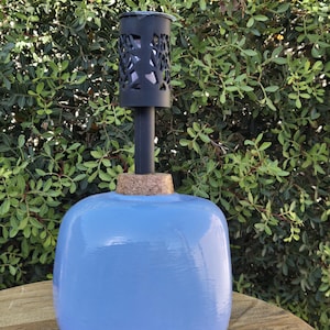 May include: A light blue, rounded ceramic oil lamp with a black metal top featuring a decorative cut-out design. The lamp has a cork base and a black metal stem. The background is a green leafy bush.
